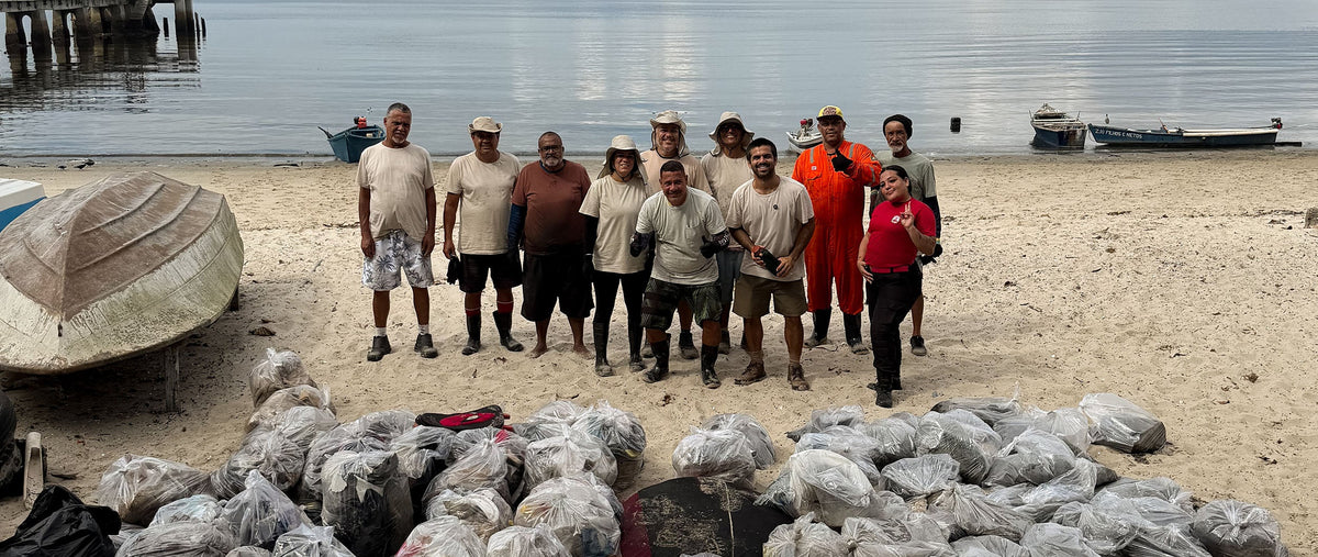 Grupo de voluntários reunido na areia após uma limpeza de praia, em frente a dezenas de sacos de lixo recolhidos, com barcos de pesca e o mar ao fundo.